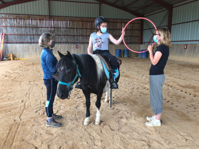 adolescent handicapé monté sur un cheval  dans le manège et faisant des exercices avec un cerceau en compagnie de marie-noëlle gachet et d'une éducatrice 