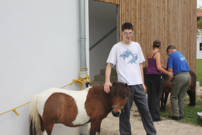 jeune homme handicapé debout caressant un petit poney ; en arrière plan un autre handicapé, de dos, caresse un autre poney, avec l'aide d'une accompagnatrice