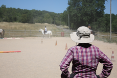 Marie Noëlle Gachet donnant un cours d'équitation dans la carrière ; elle est de dos, un chapeau sur la tête et surveille les exercices des élèves.