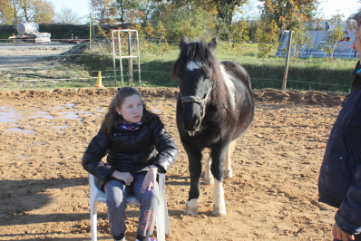 jeune fille handicapée en fauteuil ; son poney est en arrière plan et la regarde