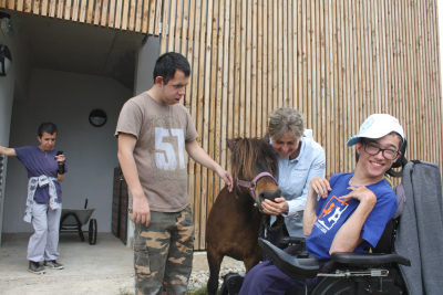 deux jeunes handicapés dont un en fauteuil roulant ont un premier contact avec un poney tenu par marie-noëlle gachet. En arrière-plan, un troisième handicapé passe un coup de téléphone.